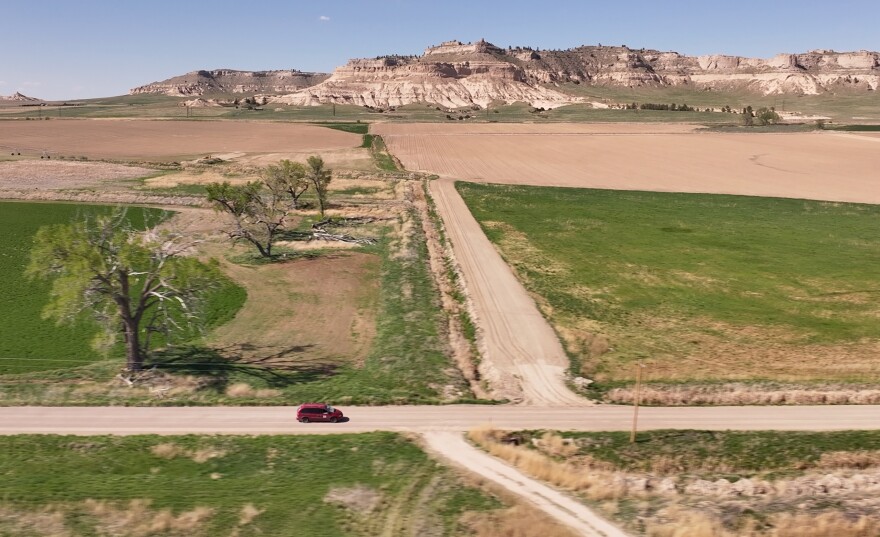 Rugged bluffs with flat tops rise toward the light blue sky in this aerial photo of northwestern Nebraska. There is little vegetation -- just a few trees and some sparse grass. A red van can be seen traveling down a road. Rural letter carrier Roger McDonald prefers to drive a Dodge Caravan on his rural route, located in Scotts Bluff County, Nebraska. The area is known for its beautiful rugged bluffs and some native prairie grasses.