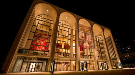 The Metropolitan Opera House facade with its distinctive arched glass front, indoor chandeliers/murals lit up at night, and the Lincoln Center plaza setting.