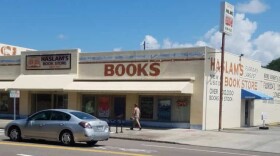 A light brown building that says Haslam's Book Store at the top and Books in large red lettering is seen from across a street with a man walking in front of it and a gray car passing to the left
