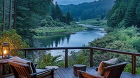 An imagined scene of a rural paradise: a wooden deck with two chairs and two foot stools overlooks a scenic river in Northern California