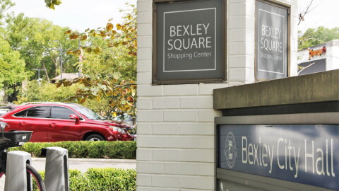 A car sits in a parking lot behind two signs reading "Bexley Square Shopping Center" and "Bexley City Hall." 