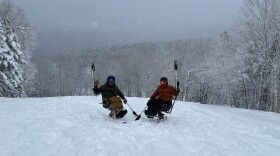 A photo of two people on adaptive mono skis next to each other on a slope, each holding up a pole, with snowy, tree covered hills in the background. Both people are wearing puffy jackets, ski helmets and ski goggles.