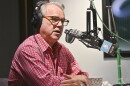 A man sitting in a radio studio sits behind a microphone and is talking.