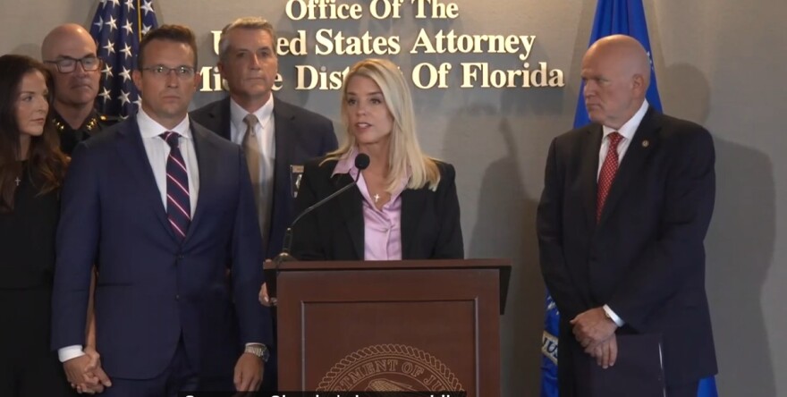 Blonde woman standing at a podium and microphone with a man in dark suit to the left, and an older bald man in a dark suit to his right