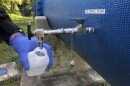 Close-up of hand with blue glove holding a clear plastic bottle under a silver nozzle with running water coming out. It's connected to a big blue pipe with the label "Lab Sample Point" on it. Greenery is in the background. 