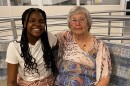 A Black teenage girl with long hair and a gray haired white woman sit together and smile