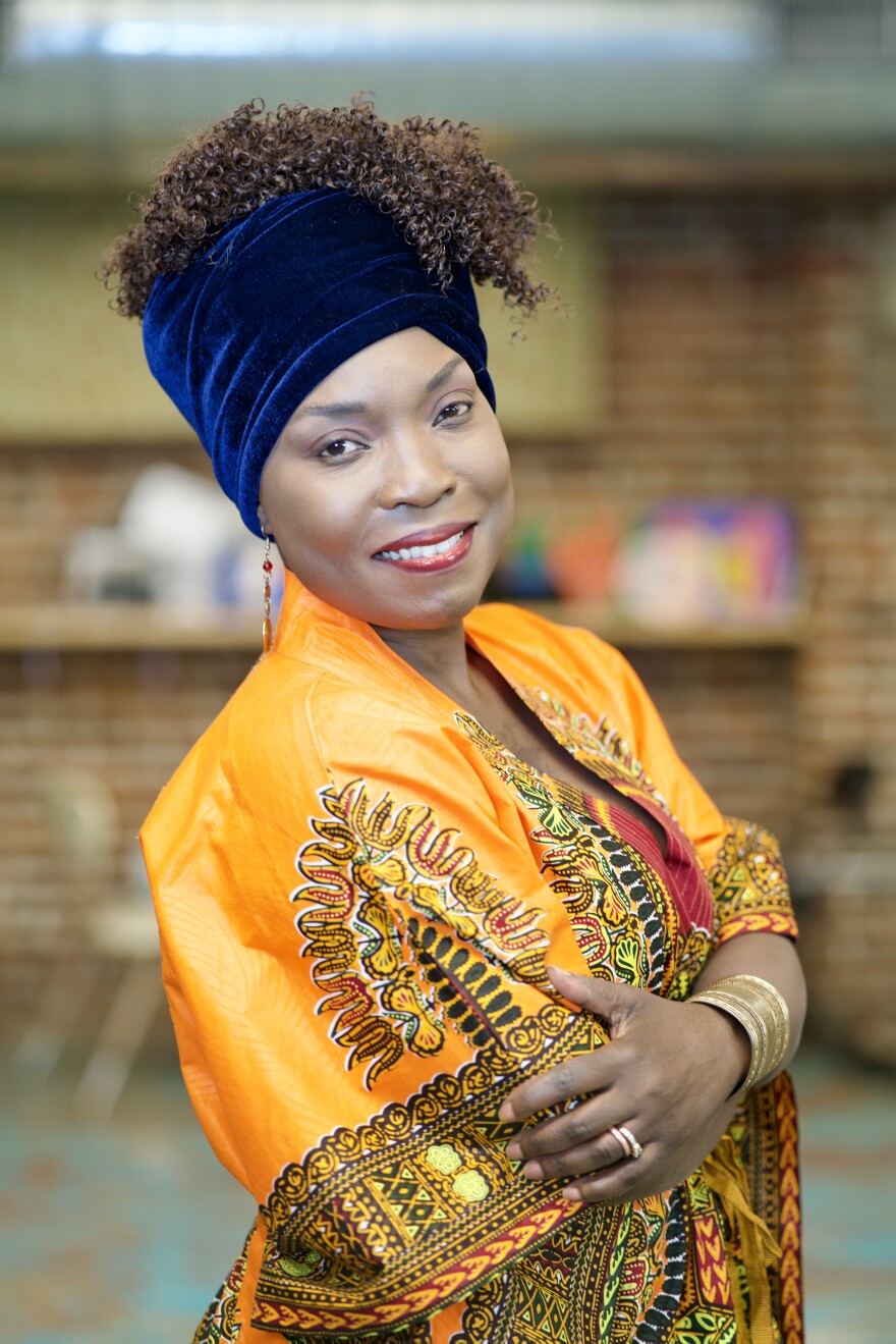 A photo of a Black woman with her hair in a velvet hair wrap, posing for a photo with her arms crossed and wearing a smile. She's wearing a bright yellow patterned scarf around her shoulders. 