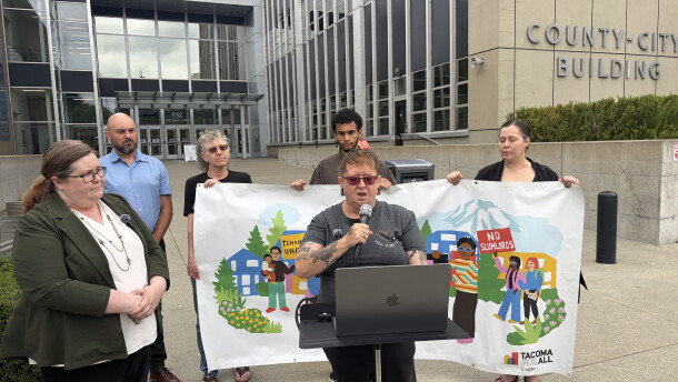 A woman speaks into a microphone while others hold a banner behind her.