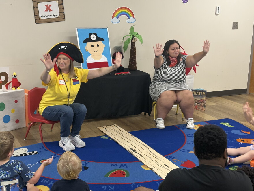Two librarians, one wearing a pirate hat, sit in chairs and hold out their hands and they lead children and dads in an activity.