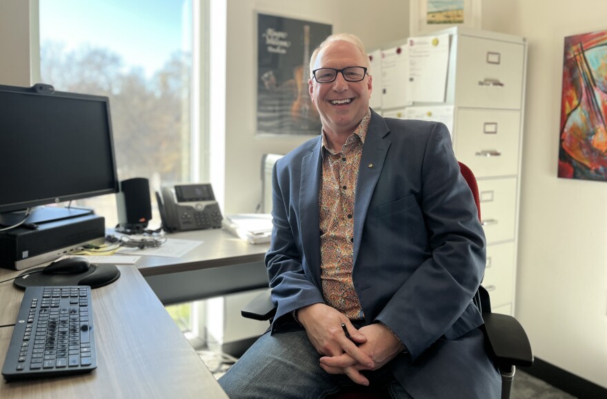 A man wearing a blazer and glasses smiles for a portrait at his office desk.