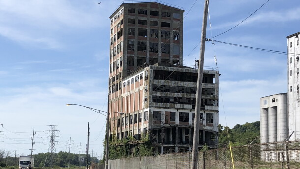 An abandoned, multi-story industrial building with broken windows stands behind a chain-link fence next to a highway.