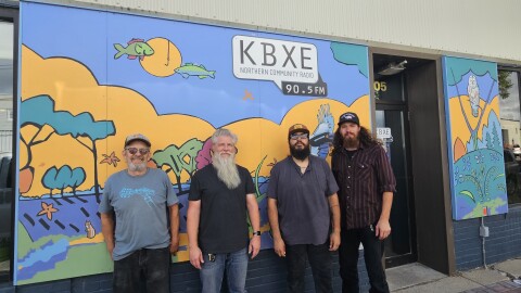 Four men standing in front of a yellow, blue, and green mural. A speech bubble over their head says "KBXE: Northern Community Radio 90.5FM"
