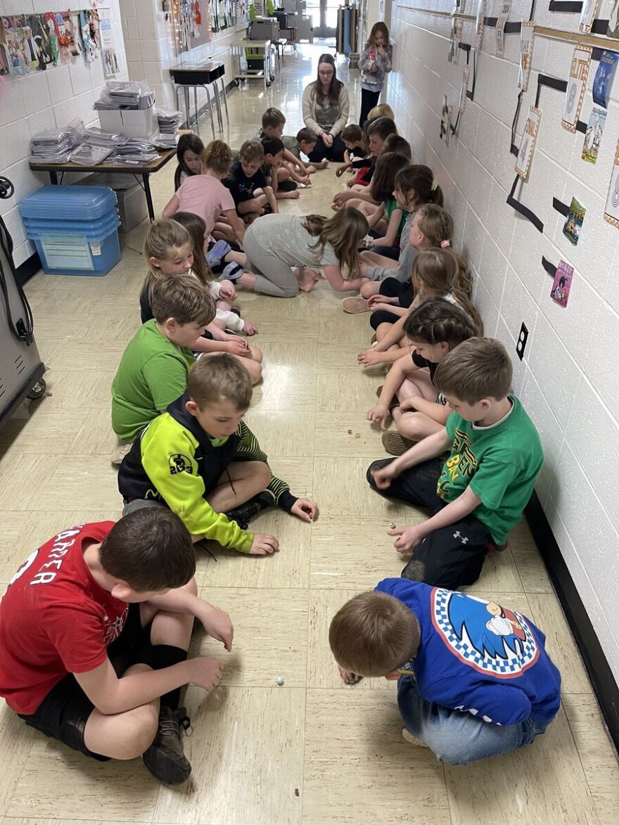 At Hunter Elementary School in the Franklin City School District, twenty-four students sit in an exterior school hallway. They are using marbles for a project. Their teacher, also sitting on the floor, observes.