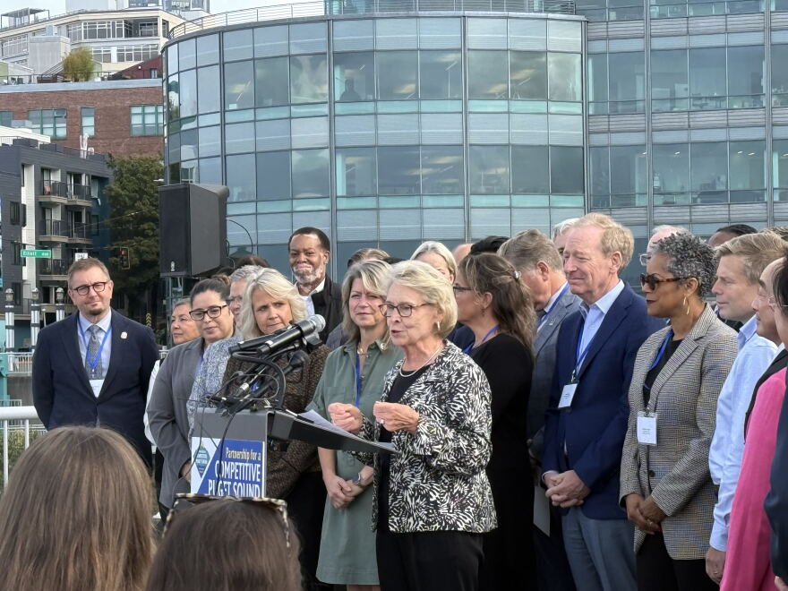 A group of civic leaders stands around former Washington governor Chris Gregoire on a rooftop. Gregoire is speaking at a podium into several microphones. 