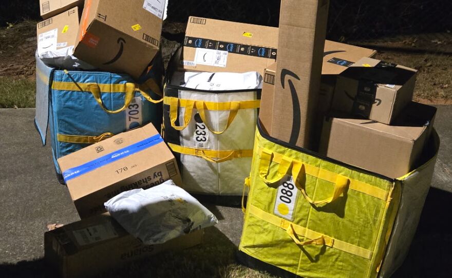 A large pile of Amazon packages and delivery bags sits on the curb at night, stacked inside and around oversized yellow and blue tote bins.