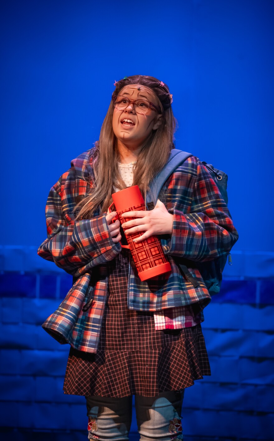 A high school actress is standing on a blue-lit stage wearing a plaid jacket and a maroon skirt is holding a canteen and wearing a blue backpack. She's singing. She has aging lines painted on her face.