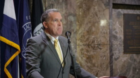 A man in a grey suit with a gold tie stands behind a podium.