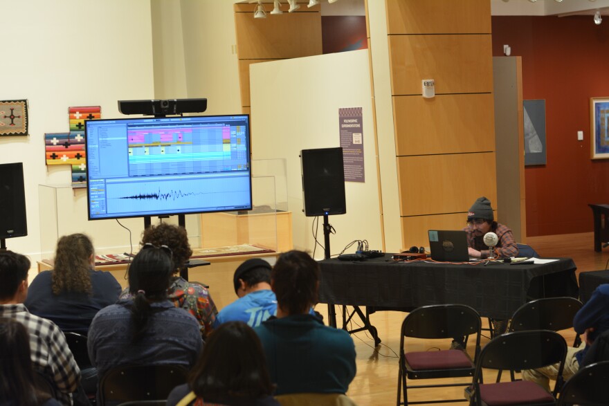 An audience watches a young Native American man perform from his laptop. Audio software is visible on a project screen next to him.