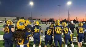 A group of football players in navy blue watch the game from the sidelines as the sun sets over the field.