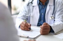 A doctor, with a stethoscope around his neck, writes on a paper on a desk
