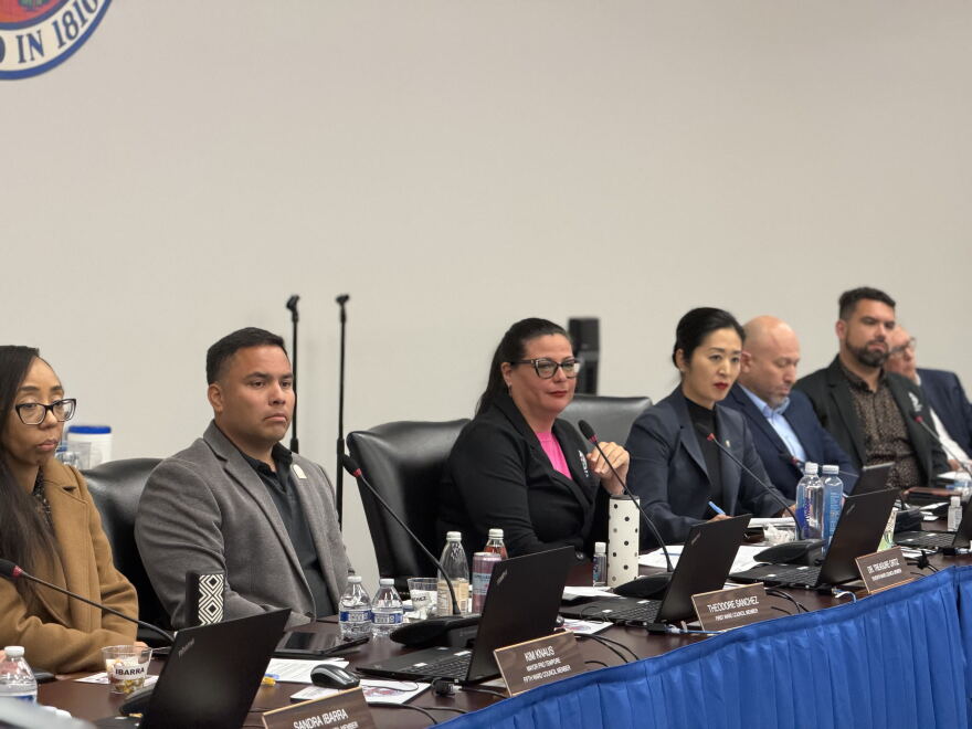 Seven councilmembers sit at a table in front of computers and microphones 