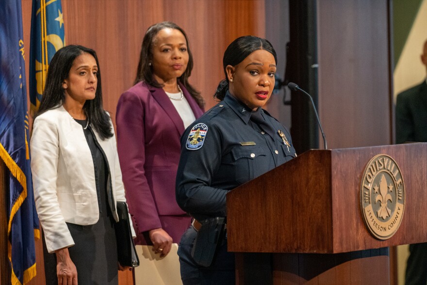 Interim police chief Jacquelyn Gwinn-Villaroel, in unifornm, speaks at a lectern. Behind her are two women.