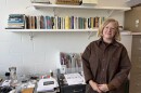 Carrie Hott in brown jacket standing in front of desk with papers and many tools and material. There is a bookshelf filled with books on the wall behind her.