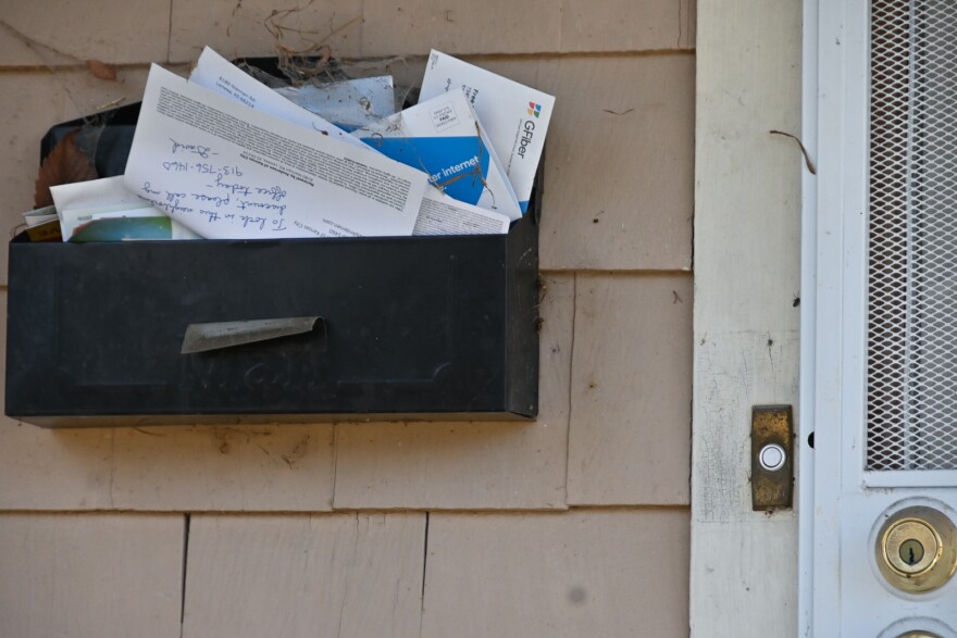 A black metal mailbox sits attached to an exterior wall of a home next to a metal door. The box is stuffed with mail that appears to have been sitting there for a while. Cobwebs can be seen stuck to some of the mail.