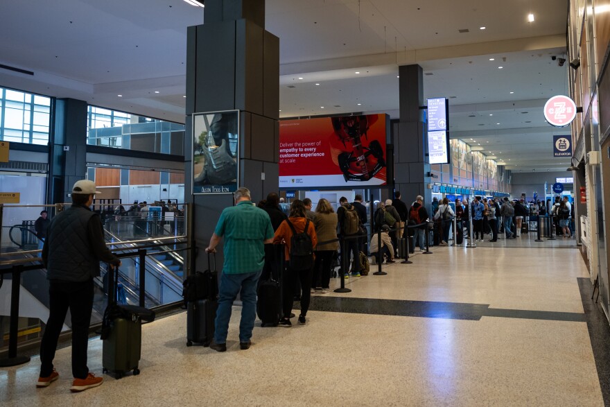 Travelers standing in line with baggage waiting to go through security. The line is long, so it wraps around a corner and you can't see the end of it. 