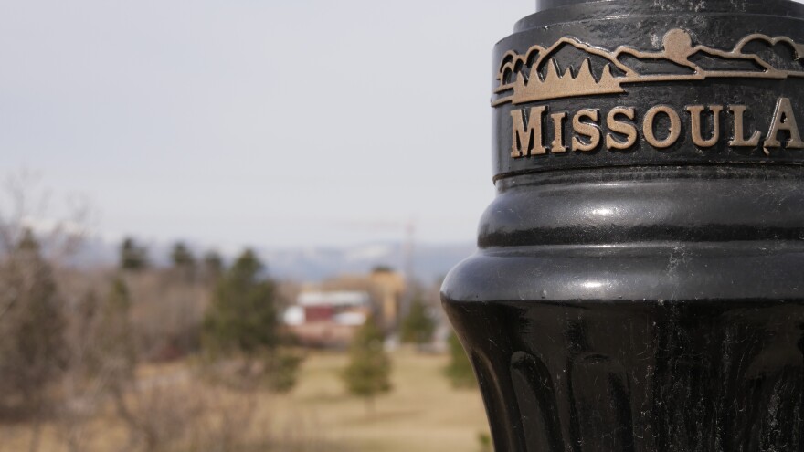 A lamp post in Missoula, MT with the word "Missoula" engraved into it. A blurred background shows a park and a skyline with buildings and mountains.