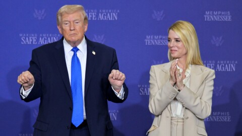 An older white man wearing a dark blur suit stands next to a white middle-aged woman with long blonde hair wearing a beige outfit. She appears to be clapping. 