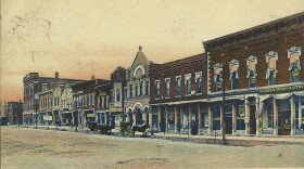 A colorized postcard showing a street view of the East Side Public Square in Bedford, Indiana during the early 1900s, featuring multi-story brick storefronts, horse-drawn carriages along the unpaved street, and text at the bottom reading "East Side Public Square, Bedford, Ind."
