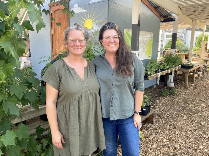 Two women smile and pose next to plants in front of a small building. Both are wearing glasses and shirts shaded green.