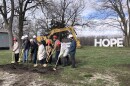 A group of men and women push gold shovels into a pile of dirt in a grass field, with trees, a construction vehicle, and large white letters spelling "hope" in the background.  