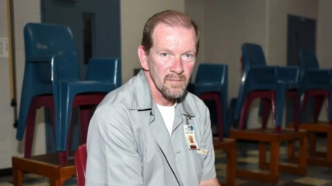 A man is sitting in a room surrounded by stacked, colorful chairs. He's wearing a gray prison jumpsuit and looking directly at the camera. 
