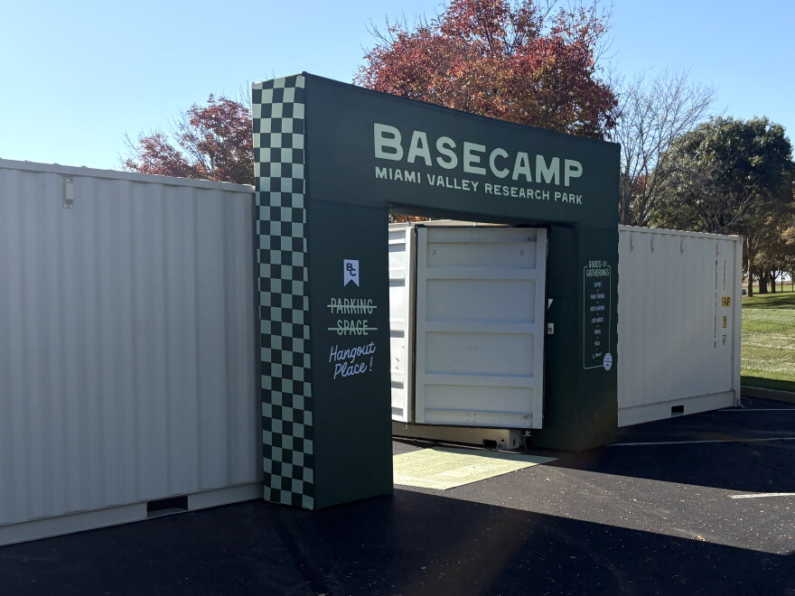 An outdoor entrance, with white shipping containers on either side. The entrance itself is dark green, which a white and green checkerboard pattern along one side. It says "BaseCamp Miami Valley Research Park" on top. Along one side, the words "Parking Space" are crossed out and replaced with "Hangout Place!"
