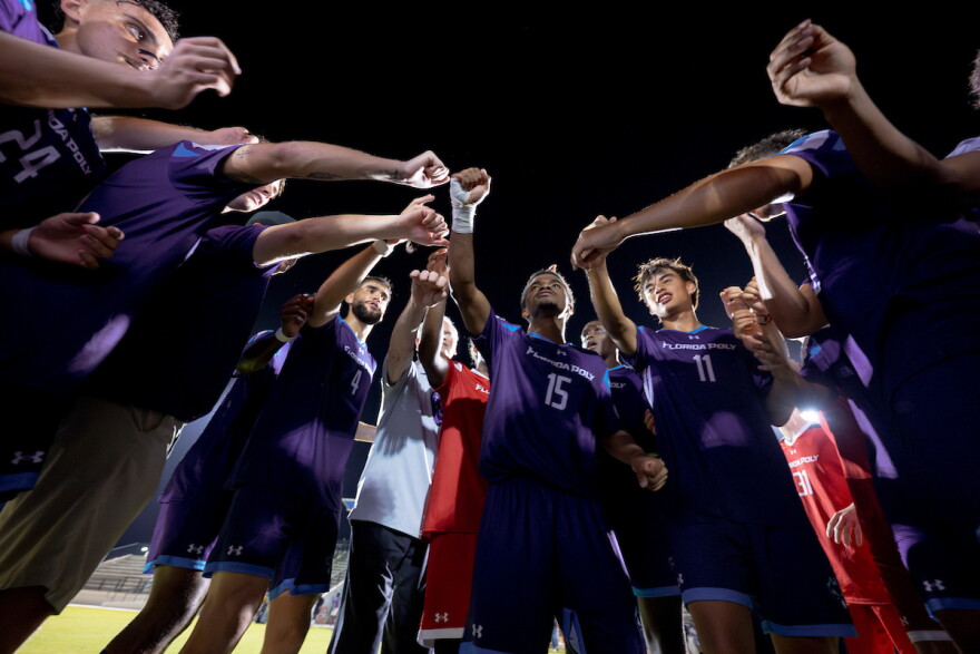 several members of a soccer team in dark blue jerseys huddle together with their arms out making a fist 