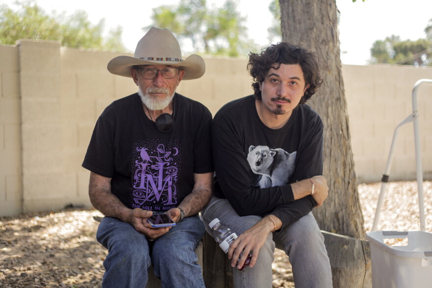 An elderly father and his adult son sit next to a tree in a backyard surrounded by a block wall. 
