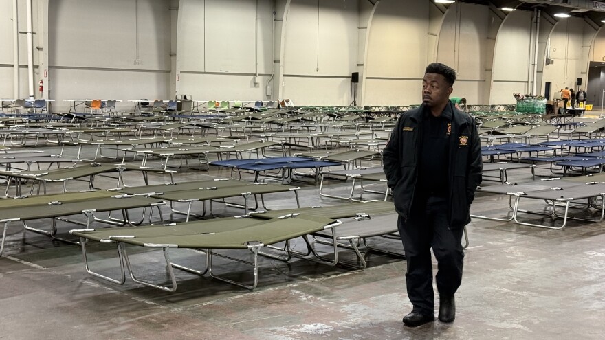 A man in a Dallas Fire-Rescue jacket walks inside a large building. Behind him are green and blue cots laid out. 