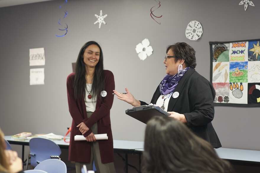 Two women stand in front of a room, speaking. One has her hand out to introduce the other. 