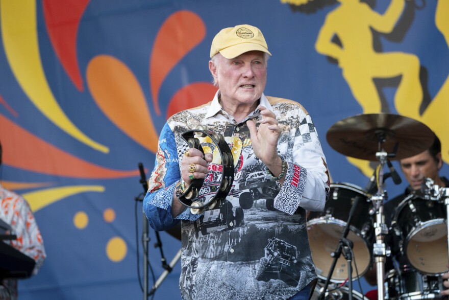 An older man wearing a colorful shirt and yellow cap plays a tambourine on stage, with a drummer and vibrant festival backdrop behind him.