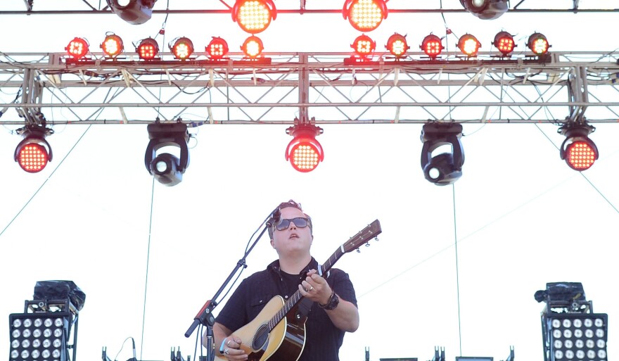 A man stands on stage holding an acoustic guitar and wearing sunglasses with a bright sky behind him and stage lights overhead.