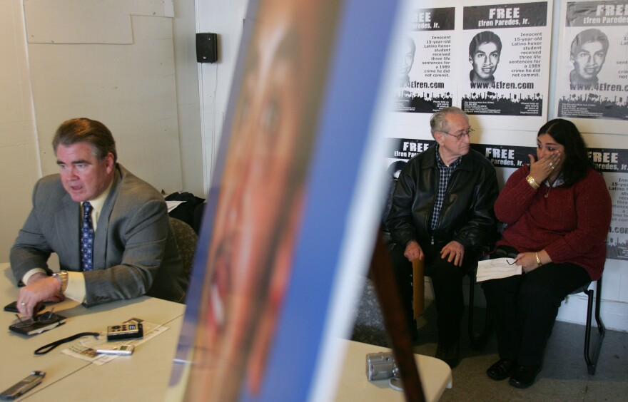 A older man sits next to a woman who is wiping tears. Behind them, posters line the wall that say "Free Efren Paredes Jr."