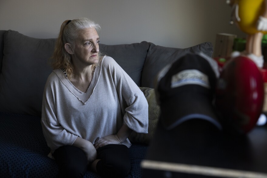 A portrait of a woman with silver-blonde hair and a light colored blouse. She has a sad, serious look on her face. She sits on a couch with natural light from a window hitting her face.