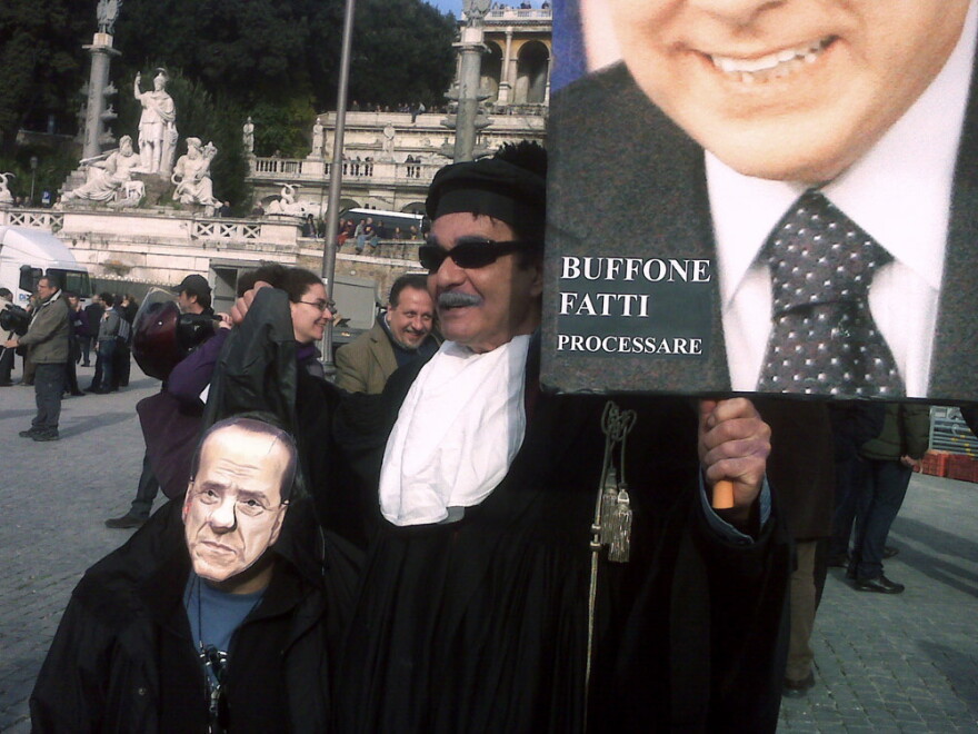 The protester to the right holds a poster calling Berlusconi a "buffoon" and prods him to show up in court.