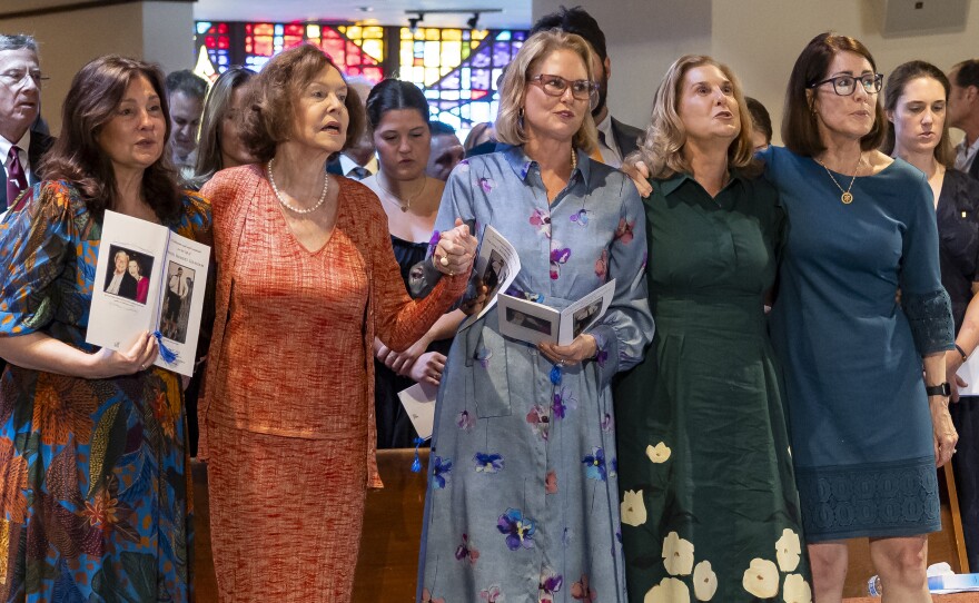 A group of women stand, holding hands, in a church. A few of them are holding programs.