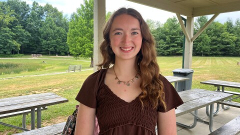 A young woman with a brown top and wavy auburn hair stands smiling in a pavilion beside picnic tables. 