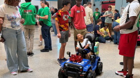 A woman stands looking at a child seated in a battery-operated toy car. The child is looking up and smiling at a man who is looking down and smiling at the child. Other people are standing an sitting in the background