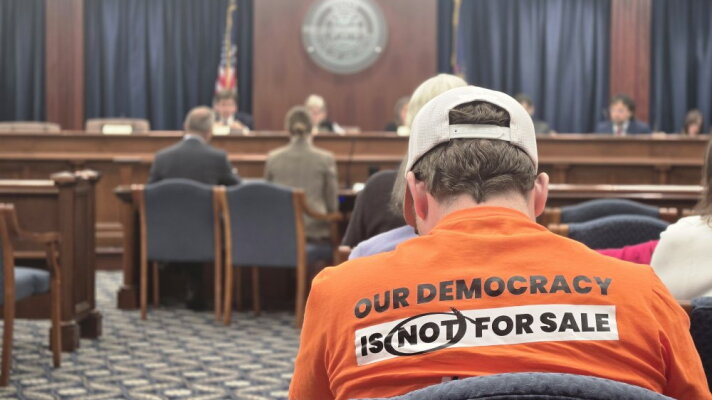 A person in an orange shirt reading "our democracy is not for sale" sits facing away from the camera as a meeting proceeds at the front of a carpeted room.