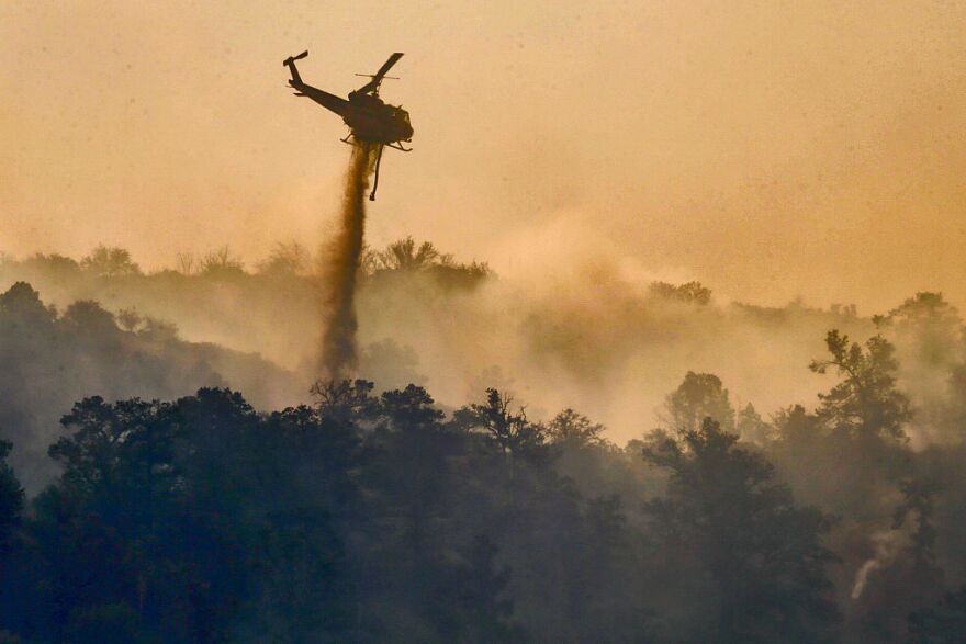 In very dense smoke from Blue Cut Fire a helicopter makes a water drop on still burning hills along Highway 2 on the way to Wrightwood.